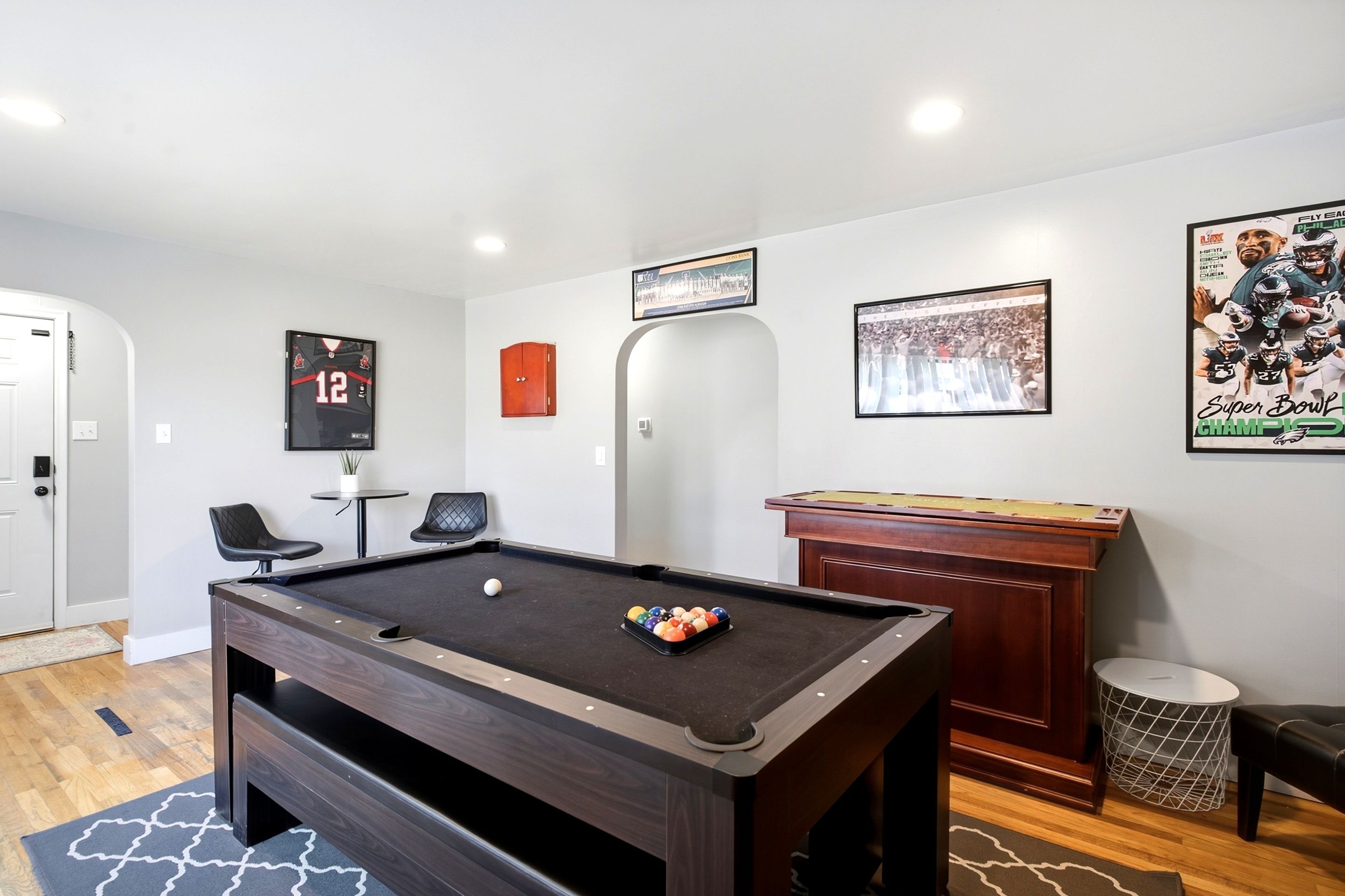 A pool table with a cue ball and billiard balls, a framed sports poster, a door, and a small round table with a plant.