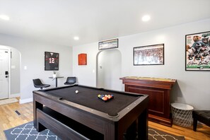 Een pooltafel met een witte bal en biljartballen, een ingelijste sportposter, een deur en een klein rond tafeltje met een plant.