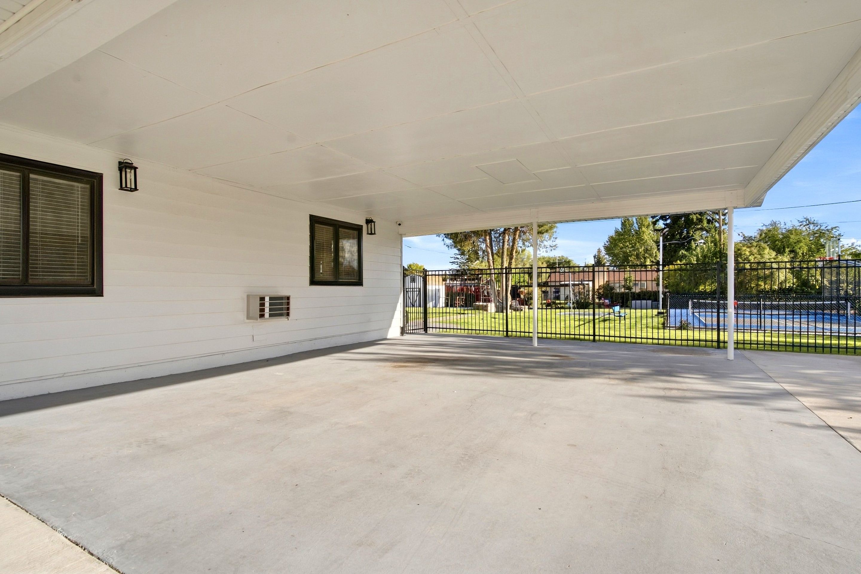 A covered outdoor area with a concrete floor, a black metal fence, and a view of a sports field in the background.