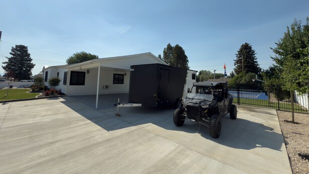 Een recreatiewagen (RV) geparkeerd op een oprit met een overdekte patio en een basketbalveld op de achtergrond.