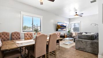 A modern living room with a dining area, featuring a wooden dining table, cushioned chairs, a grey sofa, and a flat-screen TV mounted on the wall.