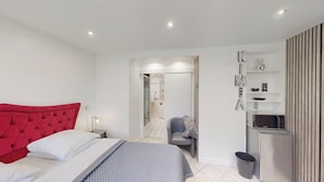 A modern bedroom with a red tufted headboard, a grey armchair, a small table, and a kitchenette area with a microwave and refrigerator. - Kimia Suite Room (Osny)