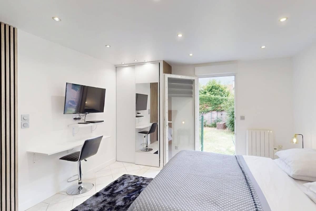 A modern bedroom with a bed, a TV, a desk, and a sliding door leading to a garden.