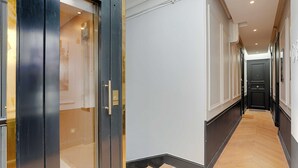 Un couloir avec une porte d’ascenseur en verre, un plancher en bois et une porte noire au fond.