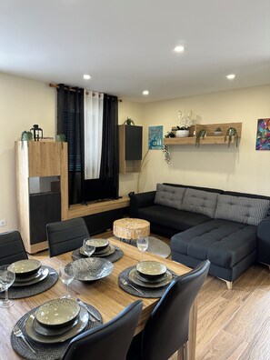 A modern living room with a dining area, featuring a wooden dining table set for four, a black sofa, and a wooden cabinet with decorative items. - Vila Romana (São Vicente, Segunda Lombada)