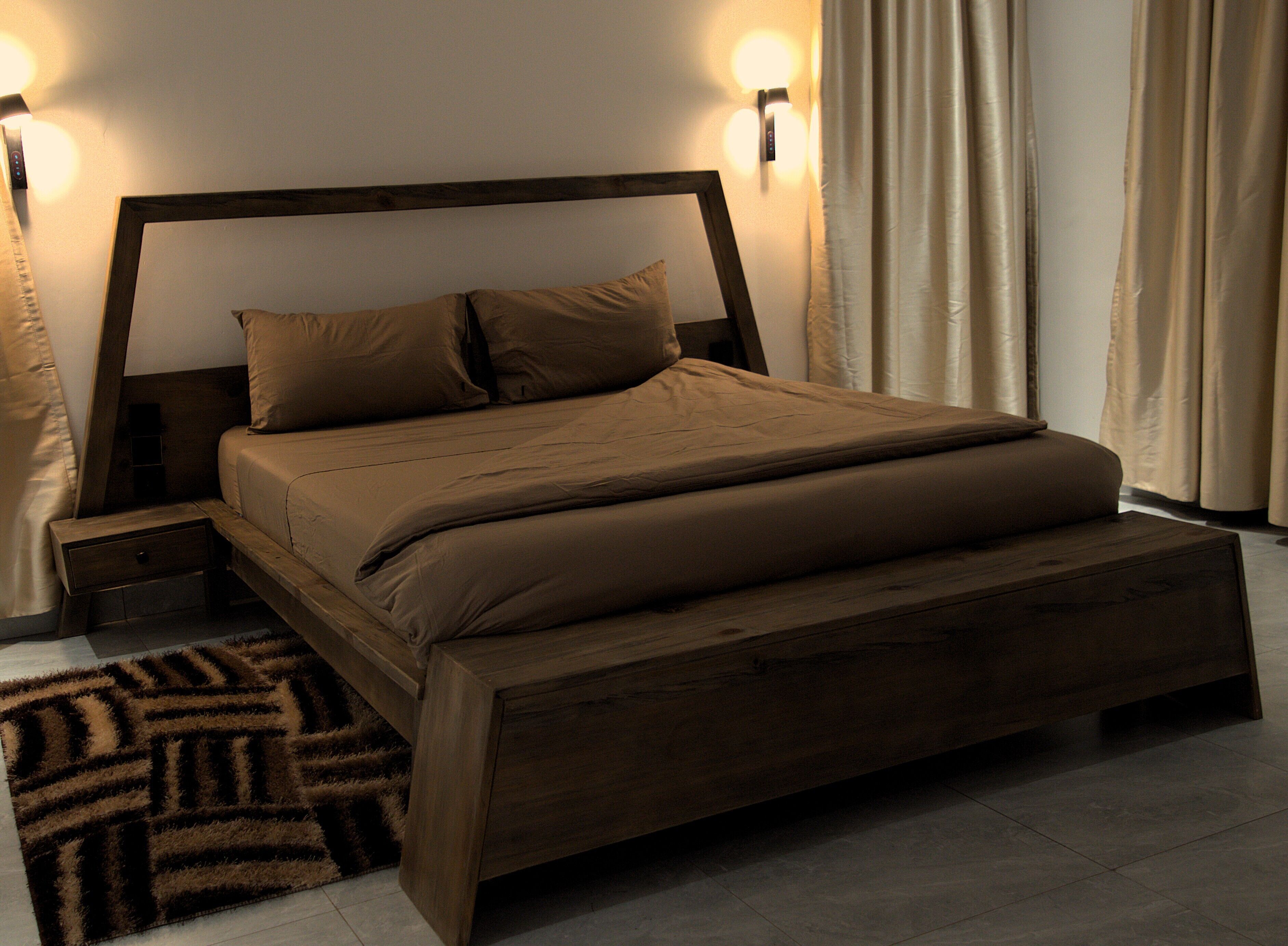 A bedroom with a wooden bed, two bedside tables, a nightstand, a patterned rug, and a wall-mounted light fixture.