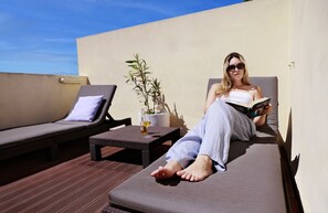 A person relaxing on a sun lounger with a book, a small table with a drink, and a potted plant.