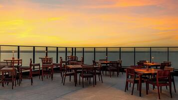 A terrace with wooden tables and chairs, overlooking a body of water during sunset.