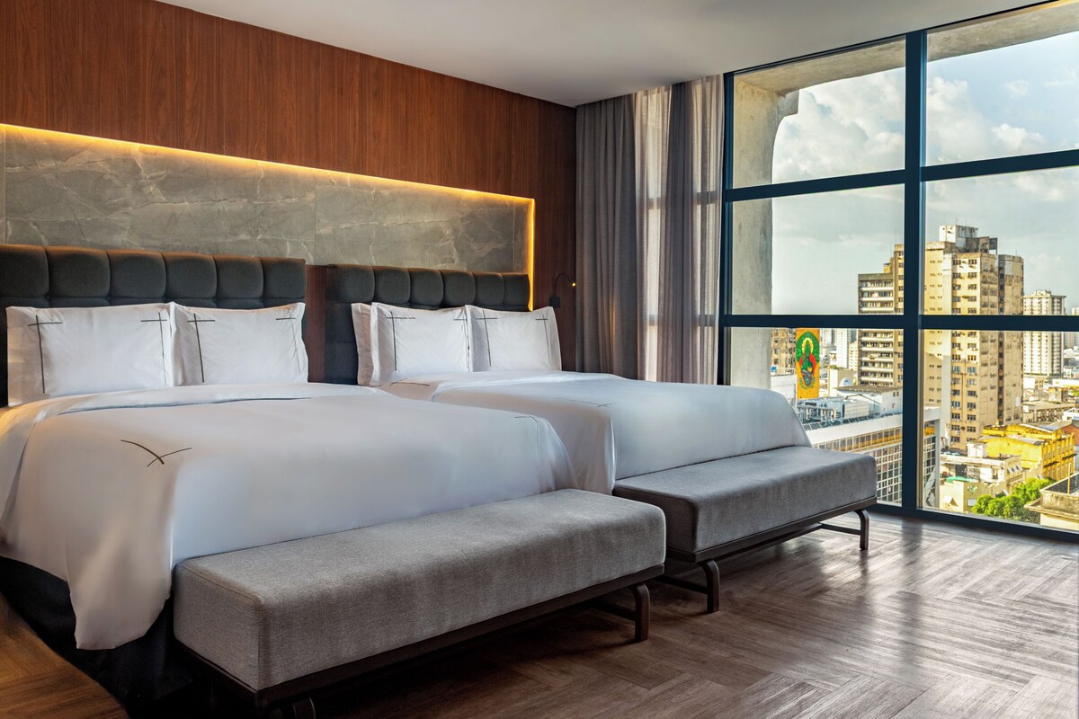 A modern hotel room with two beds, a large window with a city view, and a wooden headboard.