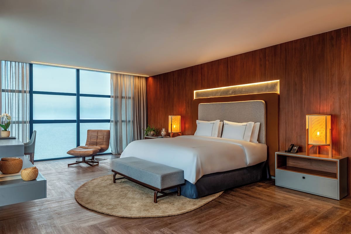 A modern hotel room with a large bed, a desk, a chair, and a view of the city through large windows.