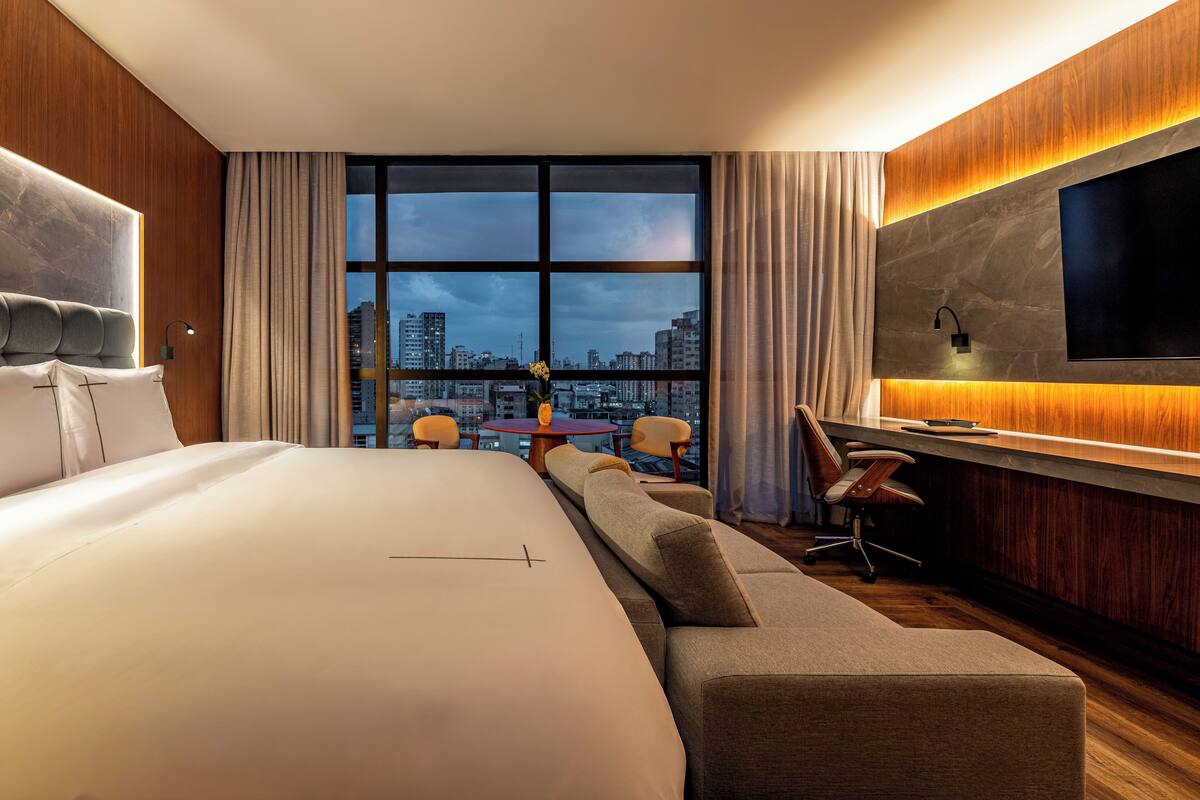 A modern hotel room with a large bed, a sofa, a desk, and a TV mounted on the wall.