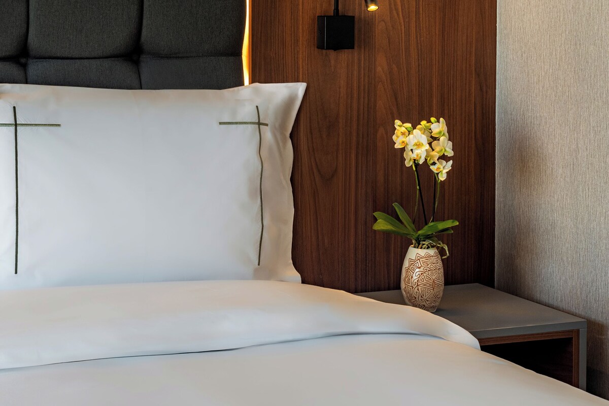 A neatly made bed with white linens, a wooden headboard, a bedside table with a vase of flowers, and a wall-mounted light fixture.
