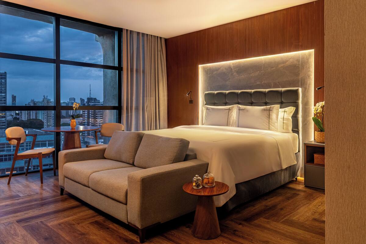 A modern hotel room with a large bed, a sofa, a round wooden side table, and a view of the cityscape through large windows.