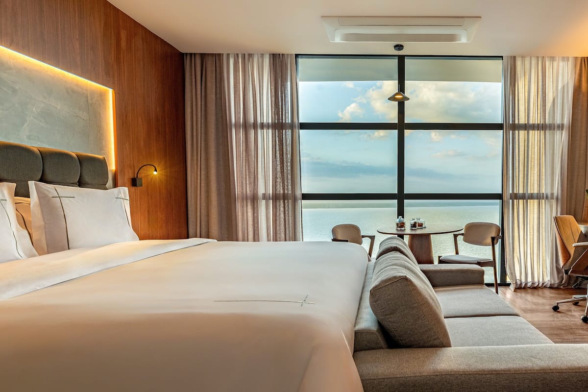 A hotel room with a large bed, a sofa, a dining table, and a view of the sea.