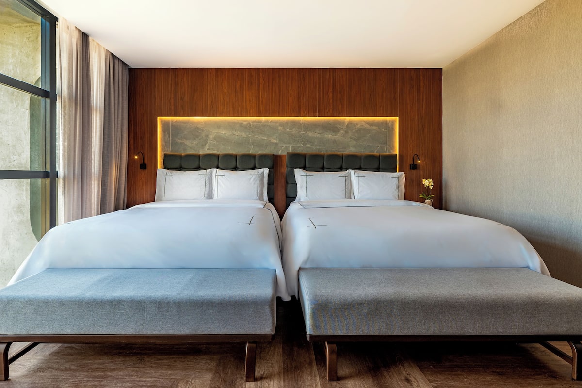 A double bed with a wooden headboard and a bedside table, a window with curtains, and a wall-mounted light fixture.