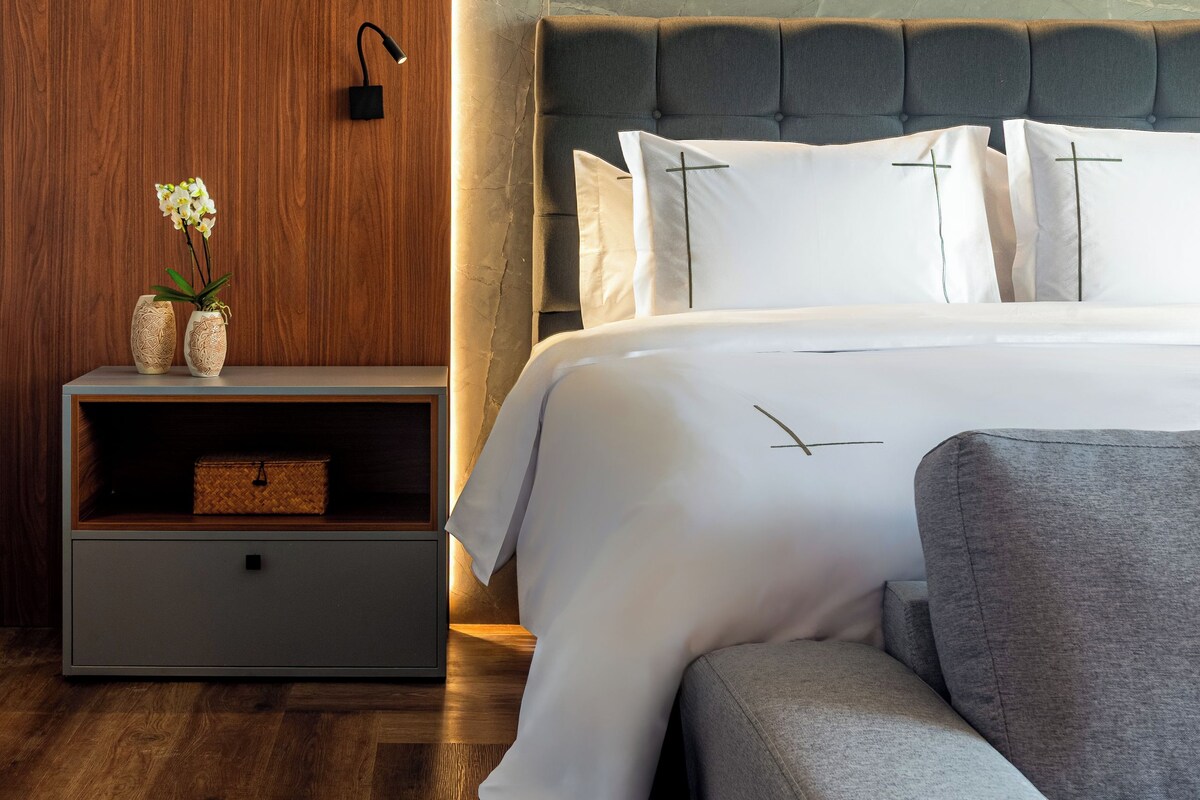 A modern bedroom with a wooden wall, a bedside table with a vase and a basket, a bed with white bedding and a grey headboard, and a grey sofa.