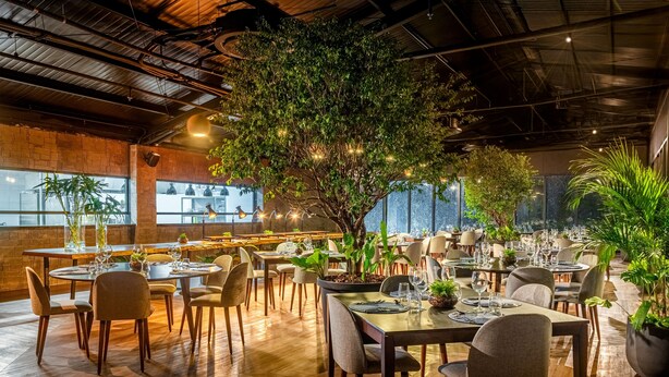 Un ristorante con diversi tavoli e sedie, un’ampia pianta interna e vista sulla città attraverso grandi finestre.