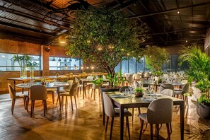 Un restaurant doté de nombreuses tables et chaises, d’un grand arbre à l’intérieur et offrant une vue sur la ville grâce à de grandes fenêtres.
