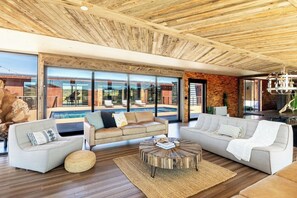 A modern living room with a pool view, wooden ceiling, and large windows. - Iconic 7,000sf Glass Villa w/ 2 Pools on 350 Acres (Pioneertown)
