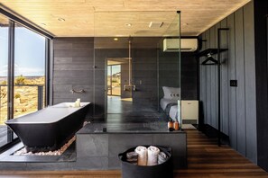 A modern bathroom with a freestanding bathtub, a glass-enclosed shower, and a large window offering a view of the desert landscape. - Iconic 7,000sf Glass Villa w/ 2 Pools on 350 Acres (Pioneertown)