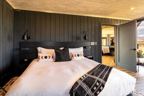 A room with a large bed, dark wooden walls, a visible door leading to another room, and a ceiling light fixture. - Iconic 7,000sf Glass Villa w/ 2 Pools on 350 Acres (Pioneertown)