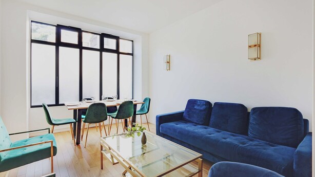 A modern living room with a blue sofa, green chairs, a glass coffee table, and a dining area with a table and chairs. - Luxurious Apartment - 3br/6p - Nation (Paris)
