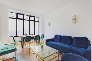 A modern living room with a blue sofa, green chairs, a glass coffee table, and a dining area with a table and chairs. - Luxurious Apartment - 3br/6p - Nation (Paris)