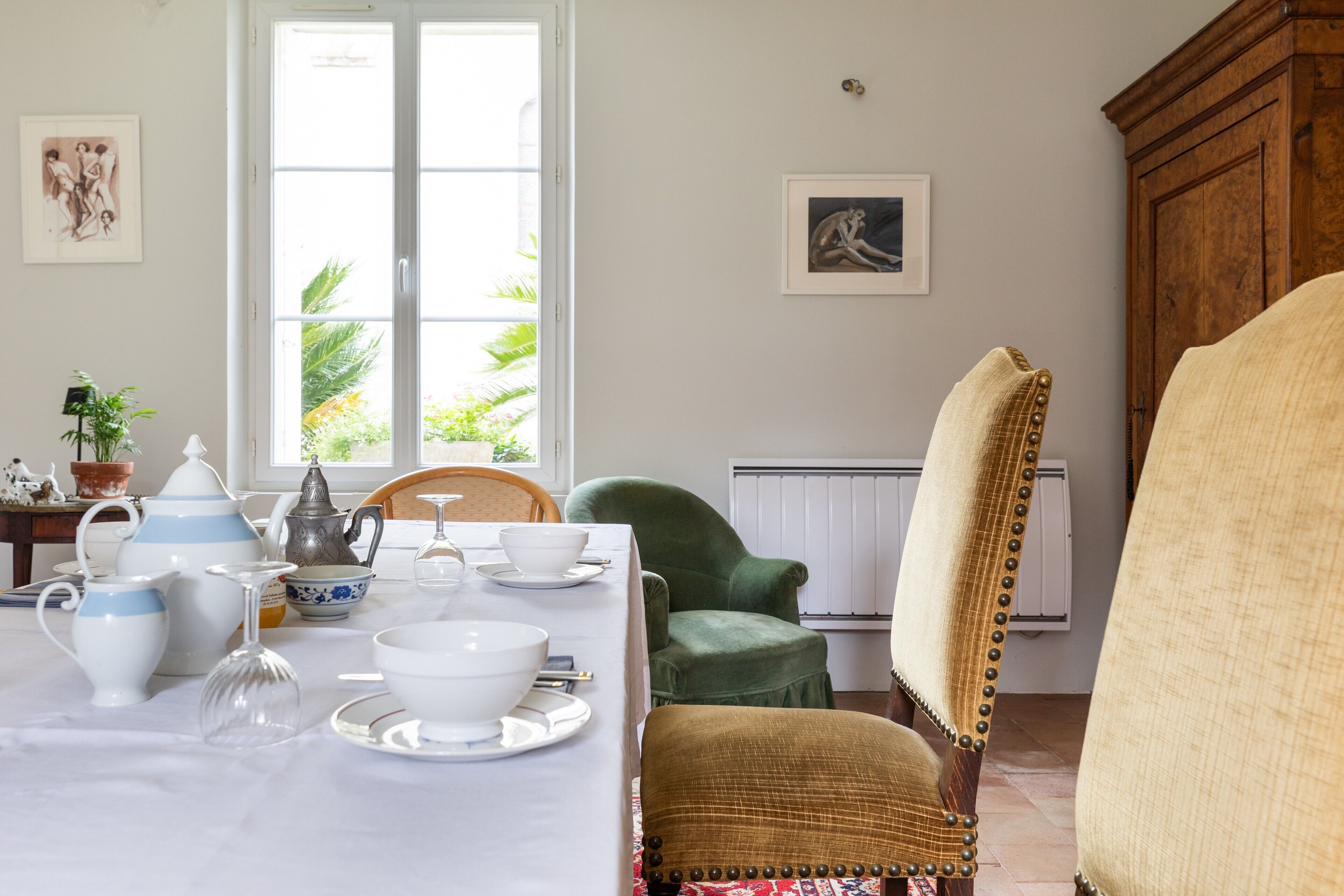 A dining area with a table set for tea, a green armchair, and a wooden cabinet.