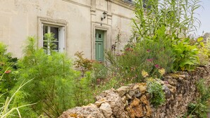 A stone wall with greenery and flowers, a building with a green door, and a window.