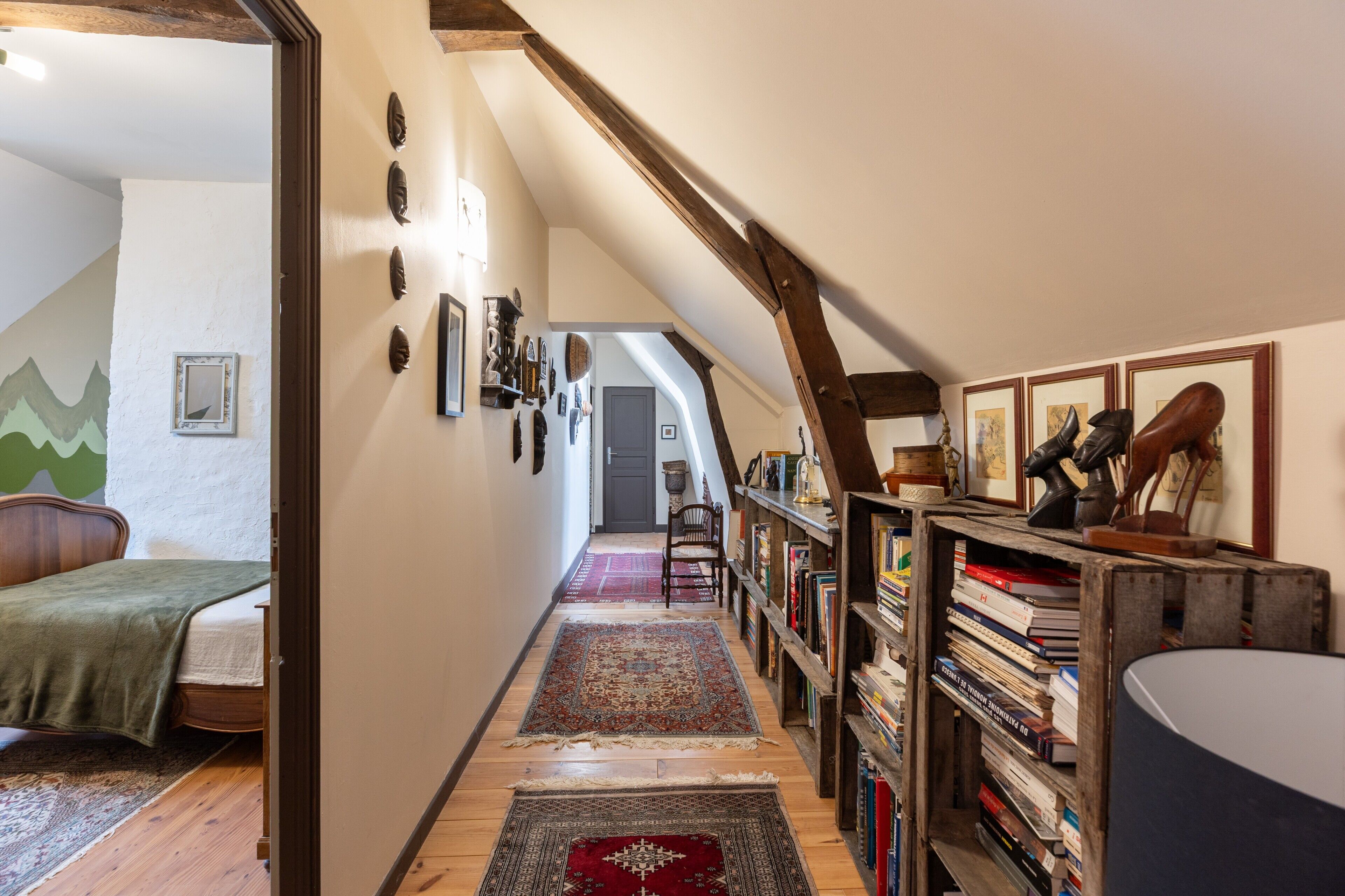 A narrow hallway with a wooden floor, a bed with a green blanket, a bookshelf filled with books, and a framed picture on the wall.