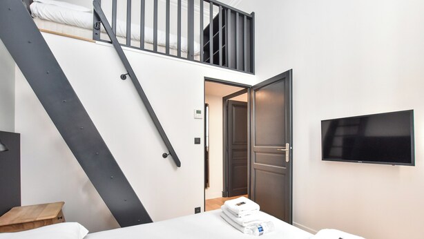 A modern hotel room with a bunk bed, a flat-screen TV mounted on the wall, and a neatly made bed with folded towels. - Charming Apartment - 2br/7p - Arc de Triomphe (Paris)