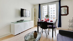 Una sala de estar moderna con una mesa de centro de vidrio, un sofá, un comedor con mesa y sillas, un televisor de pantalla plana montado en la pared y una ventana con cortinas.
