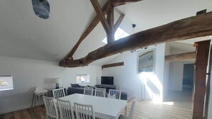 Ein geräumiger Raum mit hohen Decken, freiliegenden Holzbalken, einem Esstisch mit Stühlen, einer Couch und einem Fernseher.