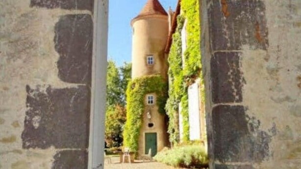 Ein rundbogiger Steingang führt zu einem Turm mit rotem Dach, umgeben von Grün.