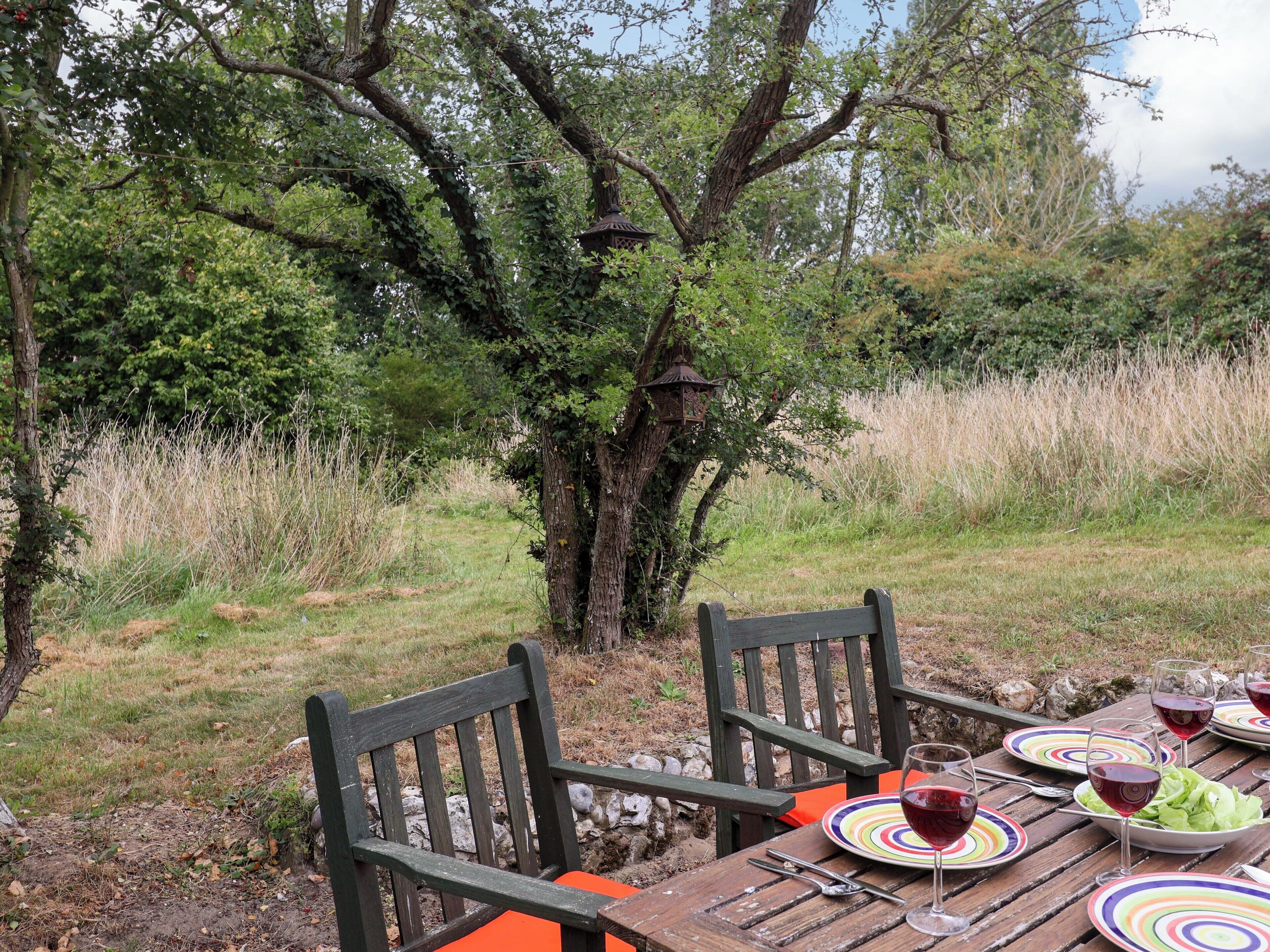 An outdoor dining area with wooden chairs and a table set for a meal, surrounded by trees and a grassy field.
