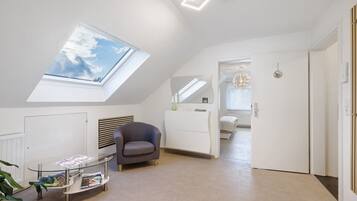 Een moderne, goed verlichte kamer met dakraam, een glazen salontafel, een grijze fauteuil en een witte garderobe.