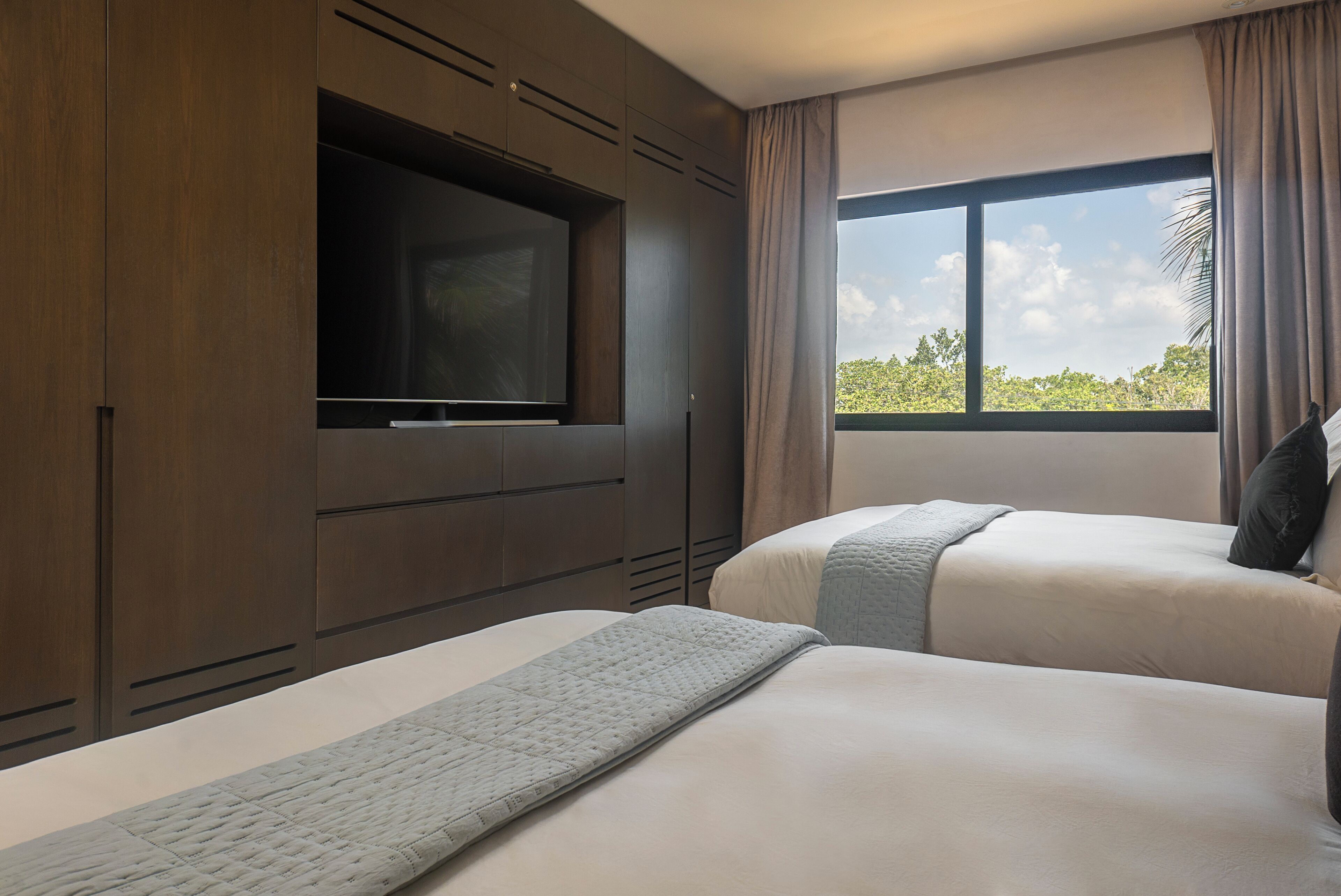 A hotel room with two beds, a TV mounted on a wooden cabinet, and a large window with a view of greenery.