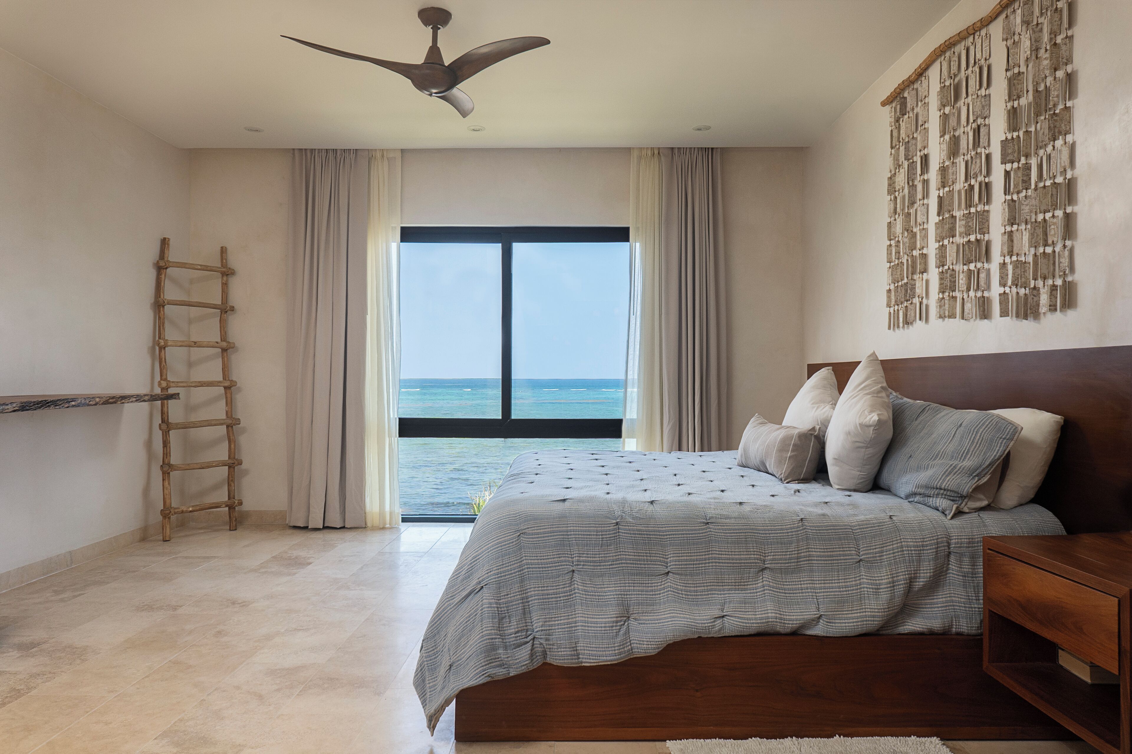 A bedroom with a large bed, a wooden ladder, a ceiling fan, and a view of the sea through a large window.
