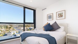 A modern bedroom with a large window, a bed with white linens, blue and gray pillows, and two framed pictures on the wall. - Bright 1BR Apartment with Stunning Views (Turner)