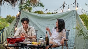 A tent with a table set for two, featuring a grill, bottles, and fruits.