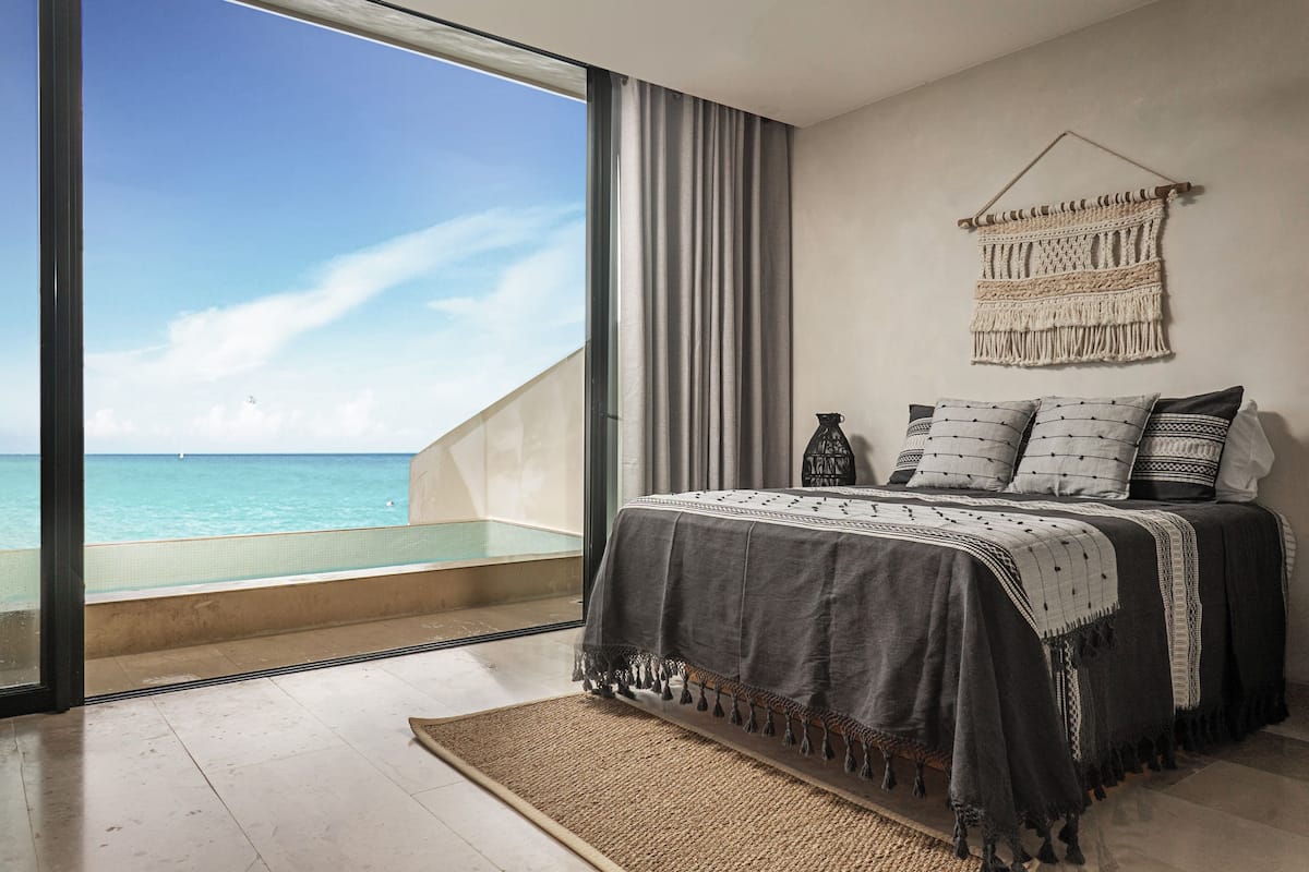 A bedroom with a large window overlooking a beach, a macrame wall hanging, and a bed with patterned pillows.