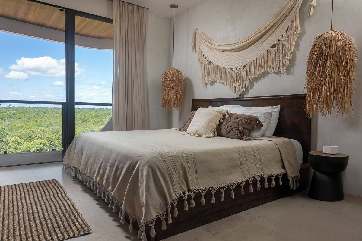 A bedroom with a large bed, a view of greenery outside, a macrame wall hanging, and a hanging light fixture.