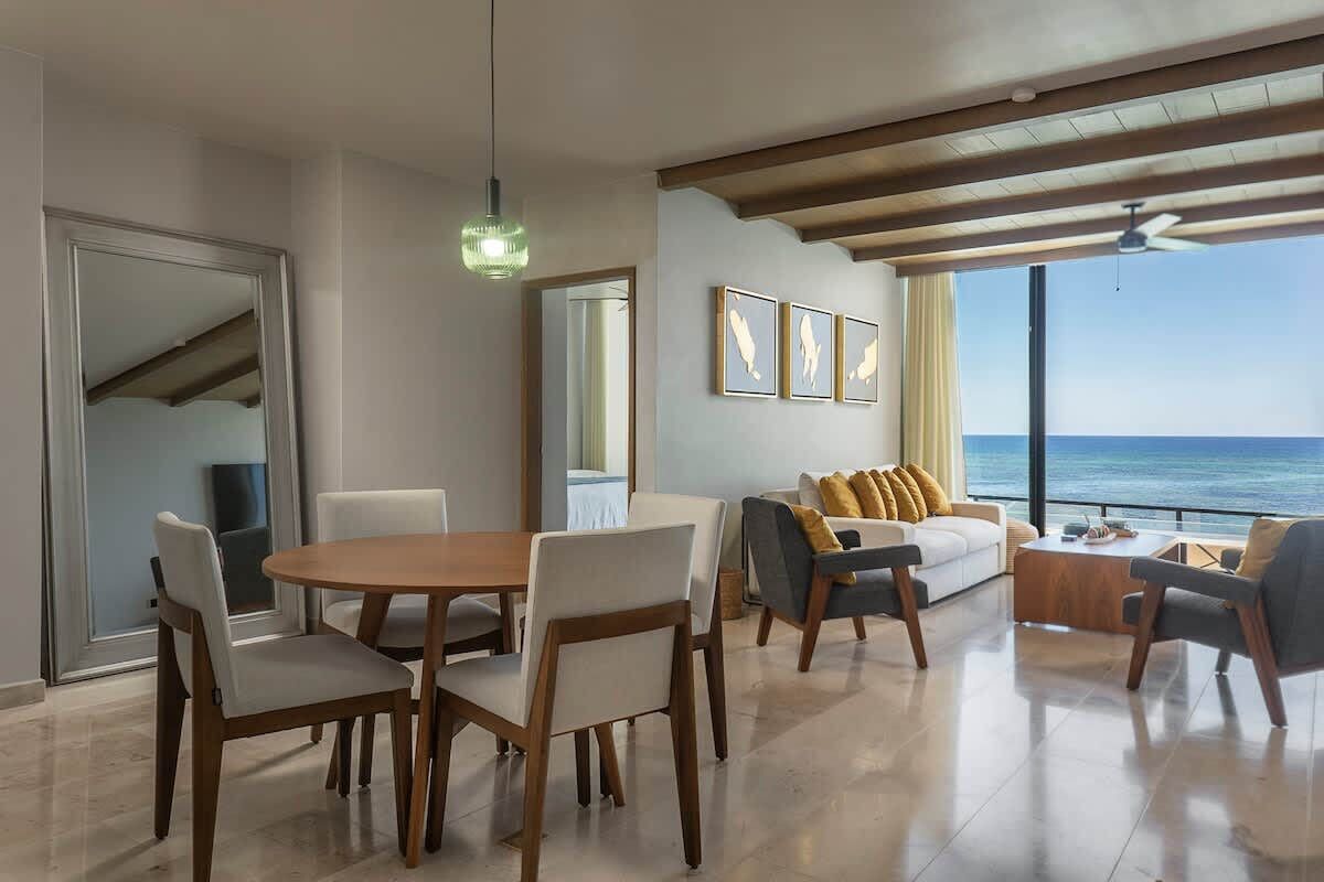 A modern living room with a dining area, large windows with a view of the ocean, and a ceiling fan.