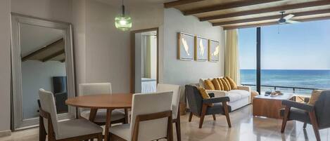 A modern living room with a dining area, large windows with a view of the ocean, and a ceiling fan.