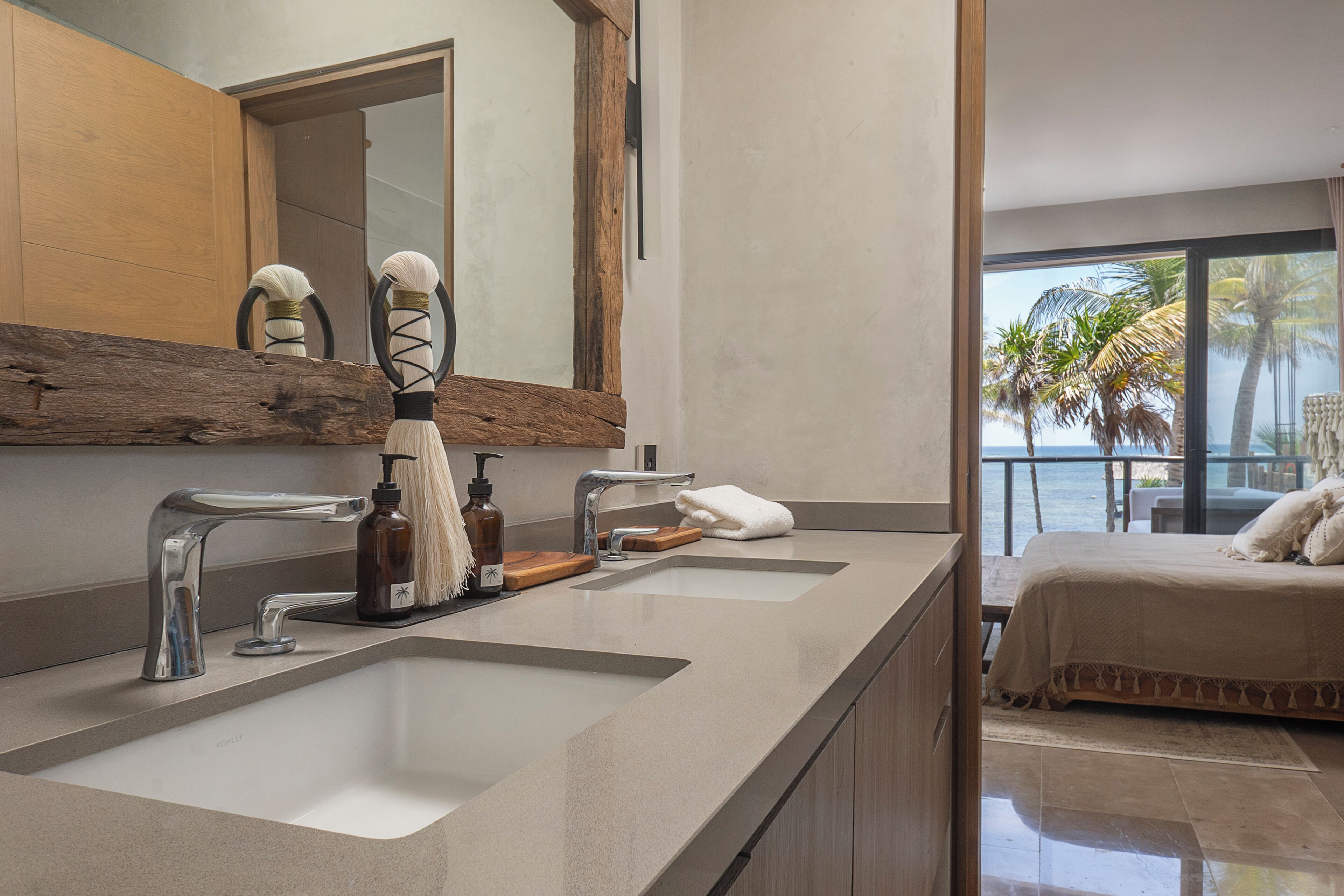 A modern bathroom with two sinks, a large mirror, and a view of a bedroom with a bed and palm trees.