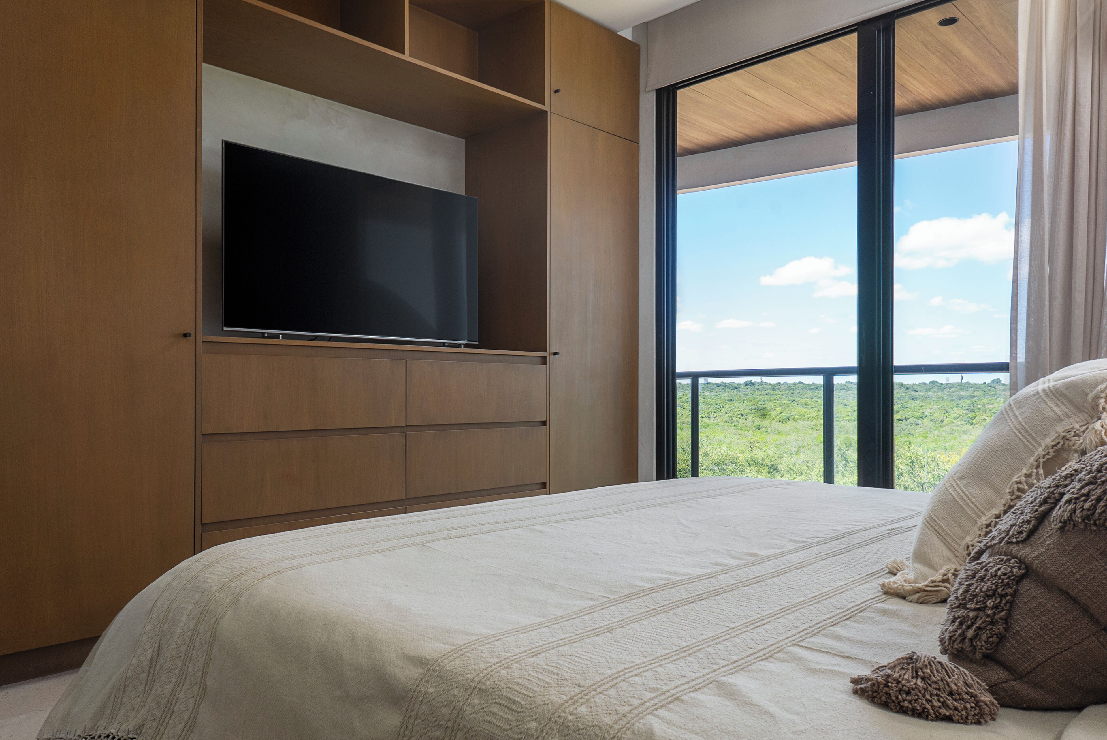 A bedroom with a large bed, a television mounted on a wooden cabinet, and a balcony with a view of the landscape.