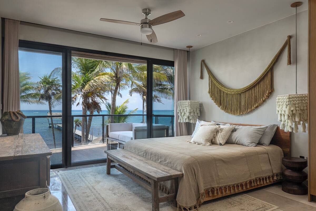 A bedroom with a large bed, a bench, a desk, and a view of the ocean through large windows.