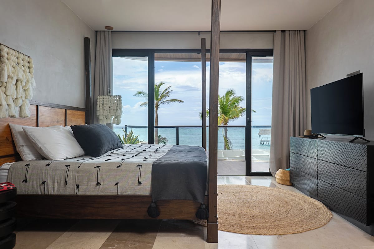 A bedroom with a bed, a TV, a large window with a view of the ocean, and a balcony.