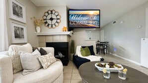 A living room with a fireplace, a television displaying "Welcome To Coastal Nest," a dining area, and a round table with decorative items.