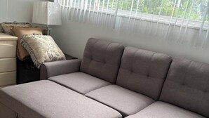 Un salon moderne avec un canapé d’angle gris, un plancher en bois et une fenêtre équipée de stores.